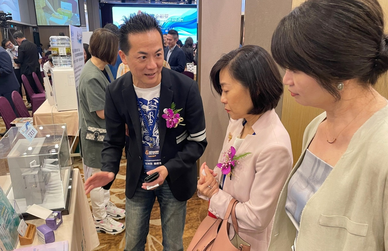 圖說：亞家生技創辦人吳坤烈博士（左）於展場攤位，向經濟部國貿署署長江文若（中）解說介紹「微型智能細胞製劑廠」。（資料來源：亞家生技）